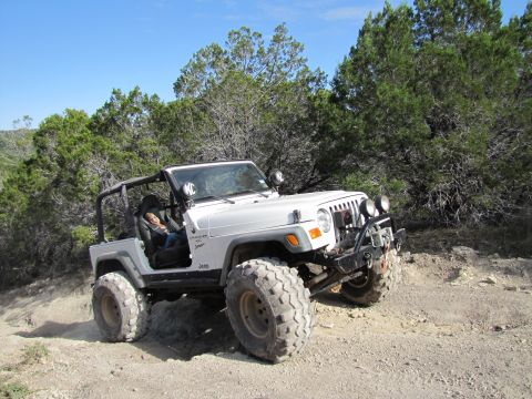 1995 Ford Bronco - A white Jeep Wrangler is parked on a dirt path surrounded by trees.