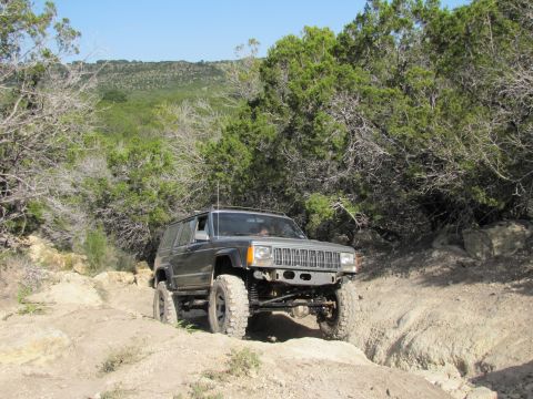 1995 Ford Bronco - Modified Jeep Cherokee XJ navigates a challenging off-road course with rocky terrain and dense vegetation.