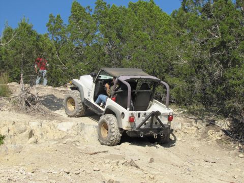 1995 Ford Bronco - White Jeep Wrangler navigates a , off-road trail with trees and rocks surrounding it.