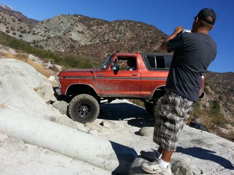 1978 Ford Bronco - A man photographs a red 1978 Ford Bronco as it drives over rocks on a sunny day.