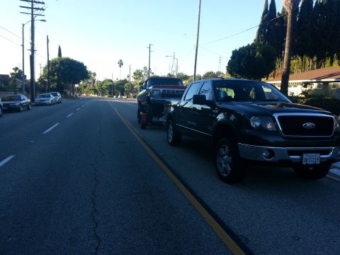 1978 Ford Bronco - A dark Ford F-150 pickup truck tows a trailer carrying another vehicle down a suburban street.