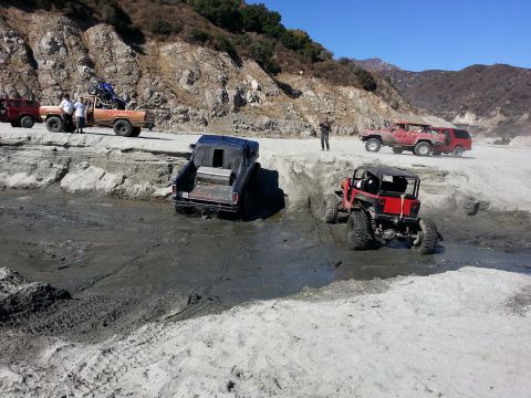 1978 Ford Bronco - Several off-road vehicles are seen navigating a challenging muddy and sandy environment, with a rocky hillside and mountains in the distanc…