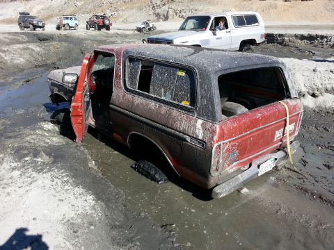 1978 Ford Bronco - Several off-road vehicles navigate a muddy terrain, with one red Ford Bronco deeply stuck.