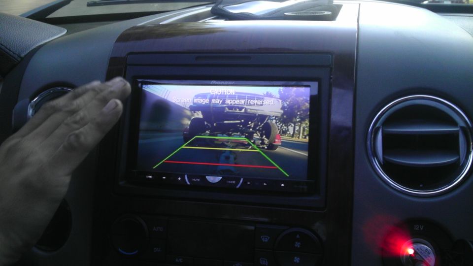 Backup camera shows a lifted truck from behind, with trees and a road visible