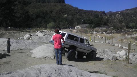 1978 Ford Bronco - A modified white SUV tackles a challenging off-road trail with large boulders and uneven terrain.