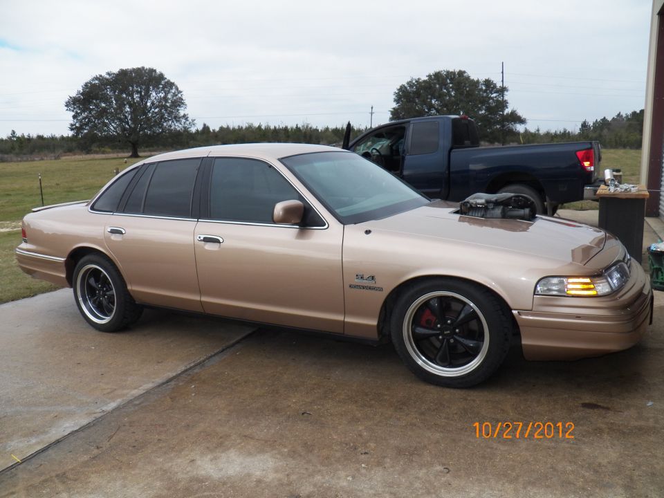 1993 Ford Crown Victoria with hood scoop and exposed supercharger, parked next to a dark pickup truck