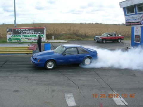 2003 Ford Crown Victoria - A blue 1987 Ford Mustang hatchback performs a burnout on a drag strip, with a red pickup truck visible in the background.
