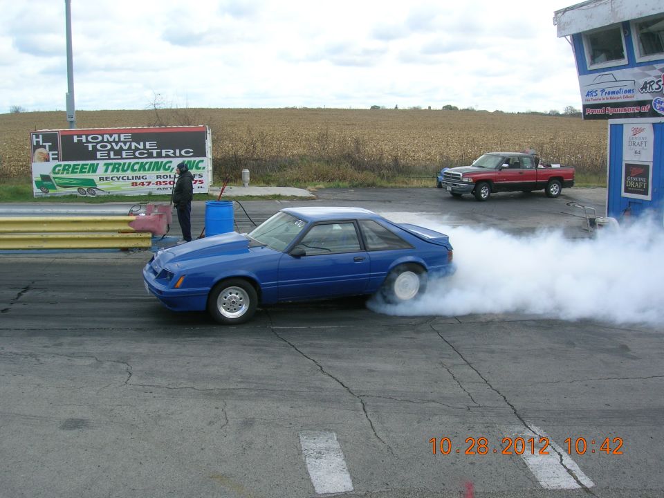 Blue 1987 Ford Mustang hatchback doing a burnout, rear tires engulfed in smoke. Aftermarket hood and wheels