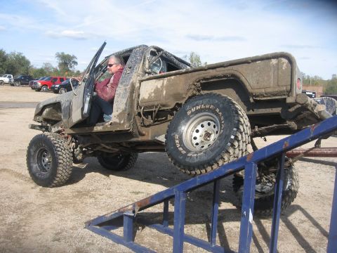 1987 Jeep Comanche - A heavily modified and mud-covered pickup truck is positioned on a ramp, with its driver visible inside.