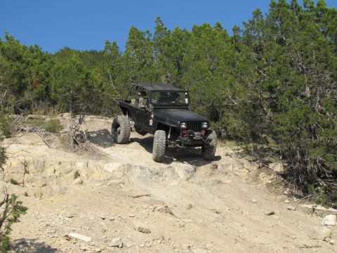 1995 Ford Bronco - A black Jeep Wrangler navigates a steep, rocky trail surrounded by dense green foliage under a clear blue sky.
