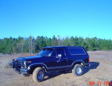 1986 Ford Bronco - Dark blue Ford Bronco parked on dirt with trees in background under a clear blue sky.