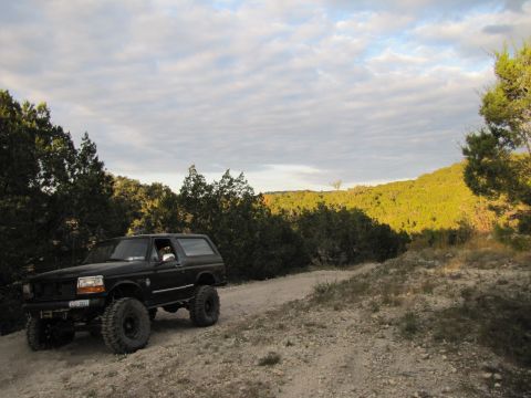 1995 Ford Bronco - A black 1995 Ford Bronco with off-road tires drives on a dusty trail through a wooded, hilly landscape.
