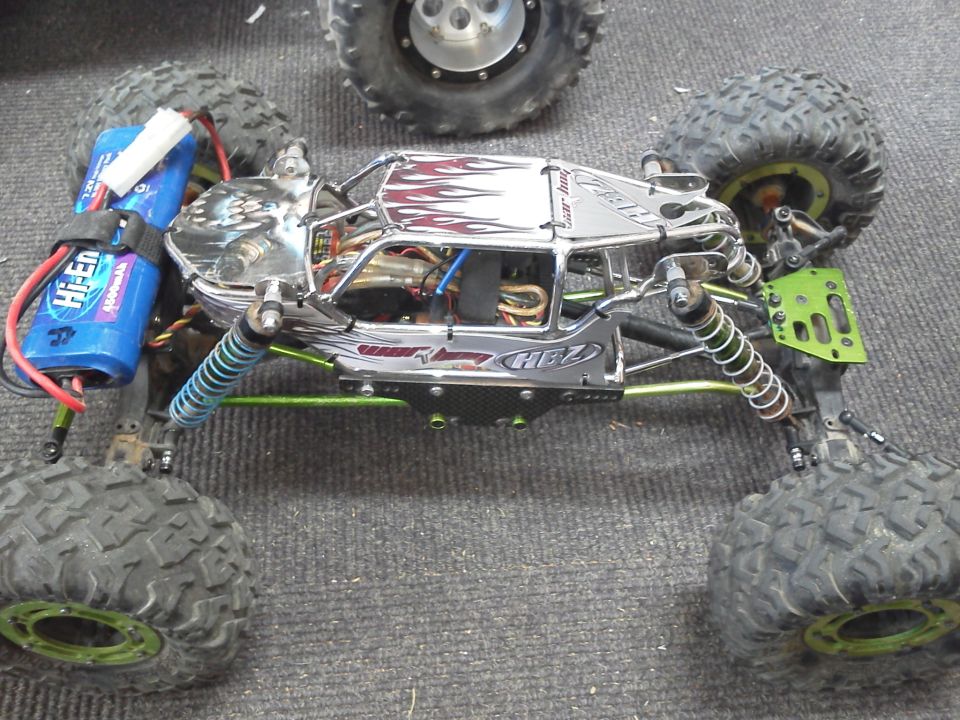 RC rock crawler with flame decals on the body, blue battery pack, and large tires. Another RC vehicle's wheel is visible in the background