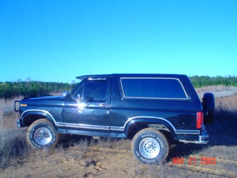 1986 Ford Bronco - Side view of a 1986 Ford Bronco parked on a dirt road under a clear blue sky.