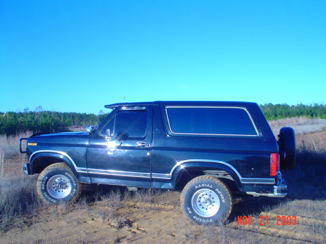 Dark blue 1986 Ford Bronco parked on dirt with trees in background