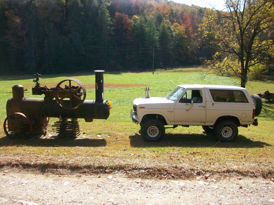 White Ford Bronco parked next to a vintage steam engine in a grassy field with trees in the background