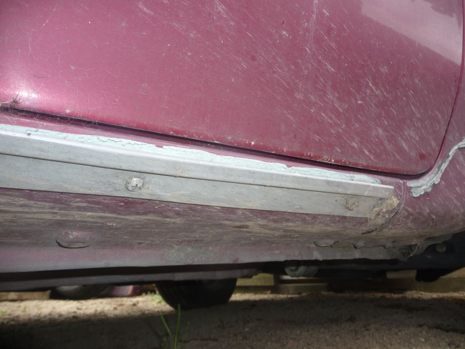 Maroon car's lower body panel with a grey trim piece attached, showing rust and damage