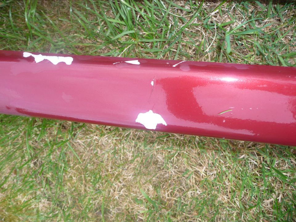 Close-up of a red car's side skirt with chipped paint, showing white primer underneath. Grass is visible in the background