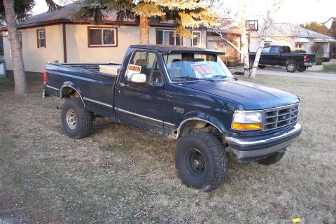 1988 Ford Bronco - A dark blue Ford F-150 pickup truck is parked in a yard with a house in the background and another truck visible.