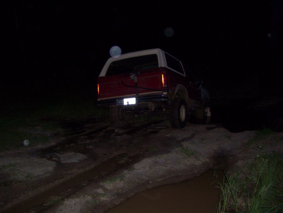 Red Ford Bronco with white top on big tires seen from behind at night on a muddy off-road trail