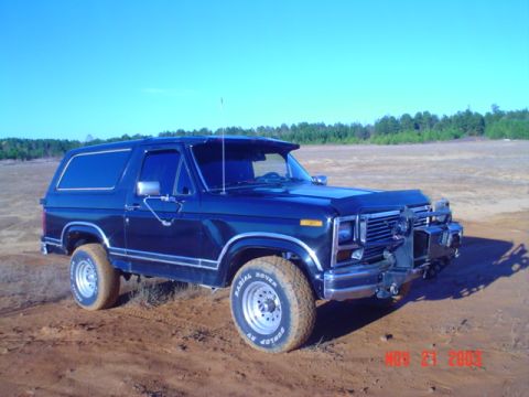 1986 Ford Bronco - This Ford Bronco is parked in a dirt lot with trees in the background.