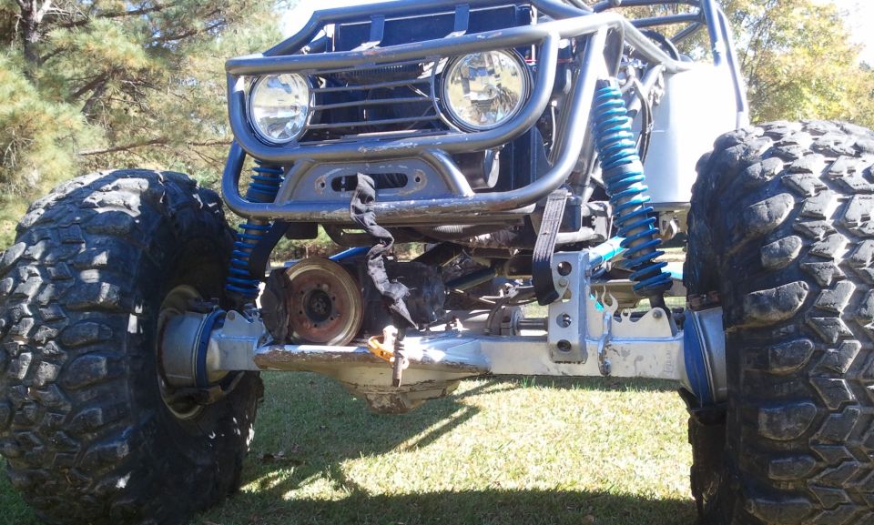 Front view of gray tube-frame rock crawler with blue coilovers, solid axle, and large mud tires on grass