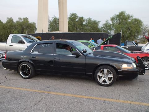 2003 Ford Crown Victoria - Lowered black full-size sedan parked among show cars near a stadium structure.