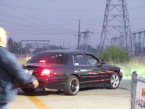 2003 Ford Crown Victoria - Dark four-door rolling toward the line at a drag event as a spectator walks past.