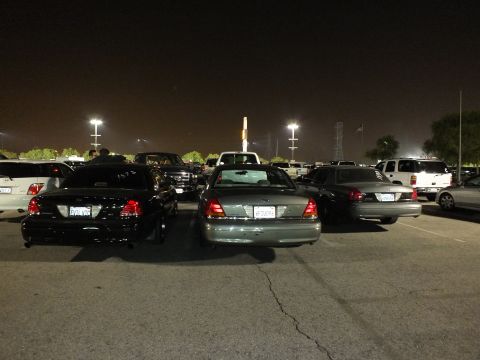 2003 Ford Crown Victoria - Black, green and silver Crown Victoria sedans angled across a dark lot under tall stadium lights