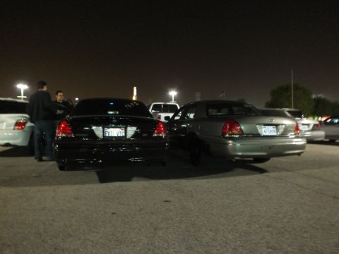 2003 Ford Crown Victoria - Two Crown Victoria rears at a night meet, black car wearing plate FYTLVTC, people standing nearby