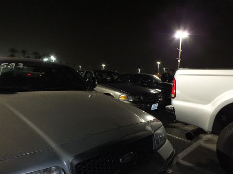2003 Ford Crown Victoria - Cluster of dark Ford vehicles and a white pickup under a single tall light pole at night