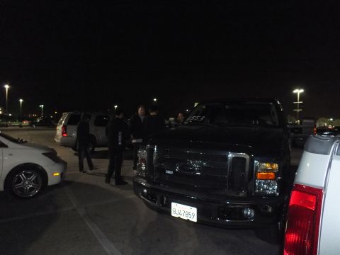 2003 Ford Crown Victoria - Black Ford Super Duty truck nose-on, plate 8J47859, people gathered beside it under lot lights