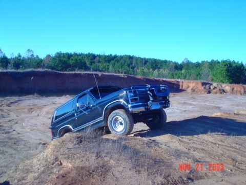 1986 Ford Bronco - A dark-colored Ford Bronco navigates a sandy, off-road terrain under a clear blue sky.