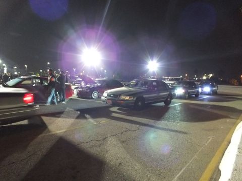 2003 Ford Crown Victoria - Crown Victoria sedans and onlookers under harsh pole lights, headlights flaring across the lot