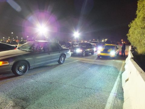 2003 Ford Crown Victoria - Vehicles lined on a night lane with headlights and pole-light glare, a yellow car among them