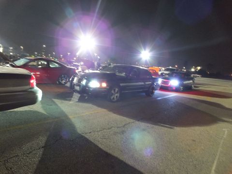 2003 Ford Crown Victoria - Black Crown Victoria headlights blazing amid parked cars under bright pole lights at night