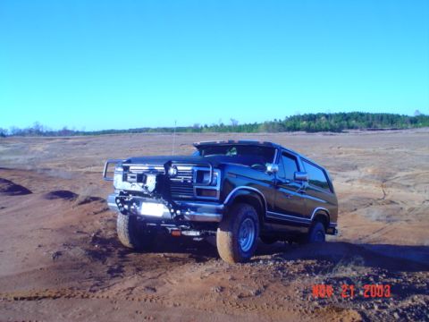 1986 Ford Bronco - A dark-colored Ford Bronco is positioned on a sandy incline under a clear blue sky.