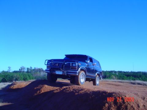 1986 Ford Bronco - A dark blue 1986 Ford Bronco is seen driving on a dirt hill with a clear blue sky above.