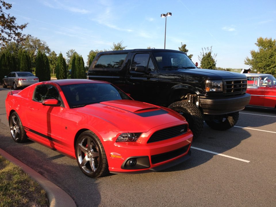 Lifted black Ford Bronco on big tires beside a red striped coupe in a parking lot.