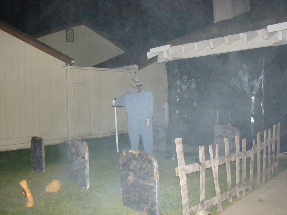 Halloween yard decorations including tombstones, a zombie figure holding a shovel, and spooky figures behind a fence