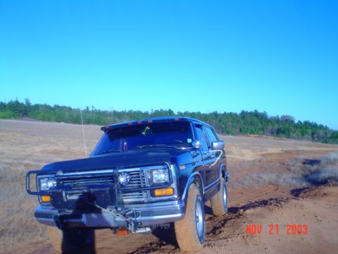 1986 Ford Bronco - A dark blue Ford Bronco navigates a muddy terrain under a clear blue sky.