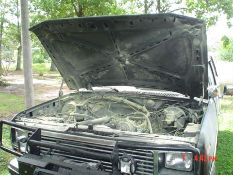 1986 Ford Bronco - The engine bay of a weathered pickup truck is exposed with the hood raised, revealing its mechanical components.