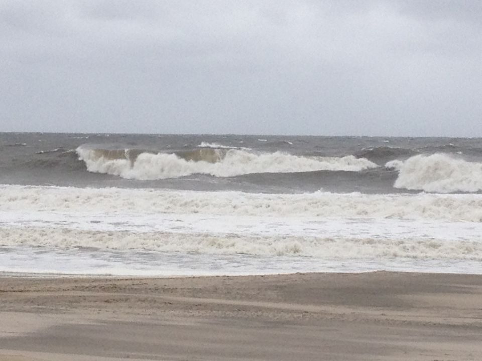 Stormy ocean waves crash on a sandy beach under an overcast sky. "SUPERMOTORS" logo visible