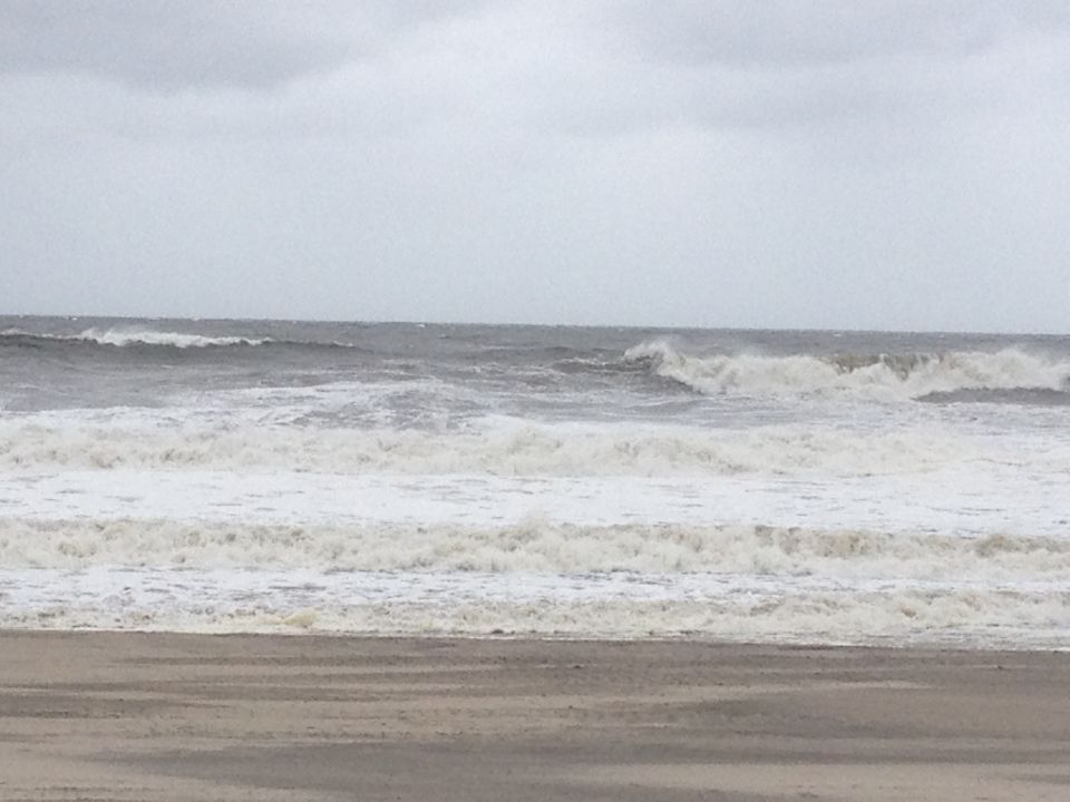 Stormy ocean waves crash on a sandy beach under an overcast sky. Watermark reads SUPERMOTORS