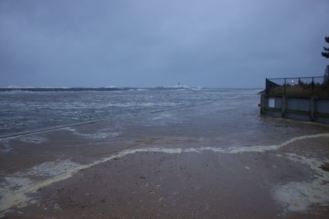 1990 Ford Bronco - A beach is inundated with ocean water and foam under a dark, overcast sky, with a fence and a distant lighthouse visible.