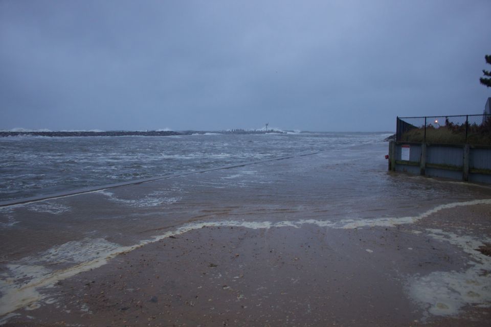 Flooded beach with foamy waves, a fence, and a distant lighthouse under a cloudy sky