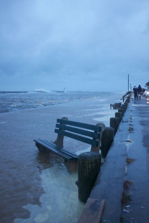 1990 Ford Bronco - A flooded boardwalk with benches partially submerged in water, as people walk along the path during a storm.
