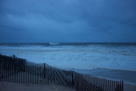1990 Ford Bronco - A fence stands on a sandy beach as large waves crash ashore under a stormy sky.