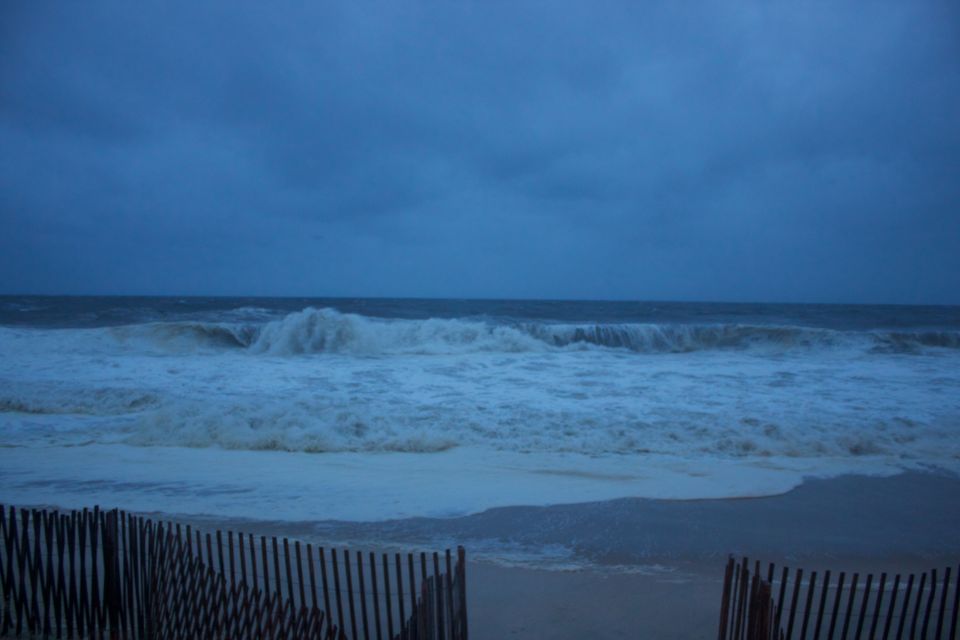 Stormy ocean waves crash on a sandy beach behind a wooden fence, with a dark, cloudy sky overhead