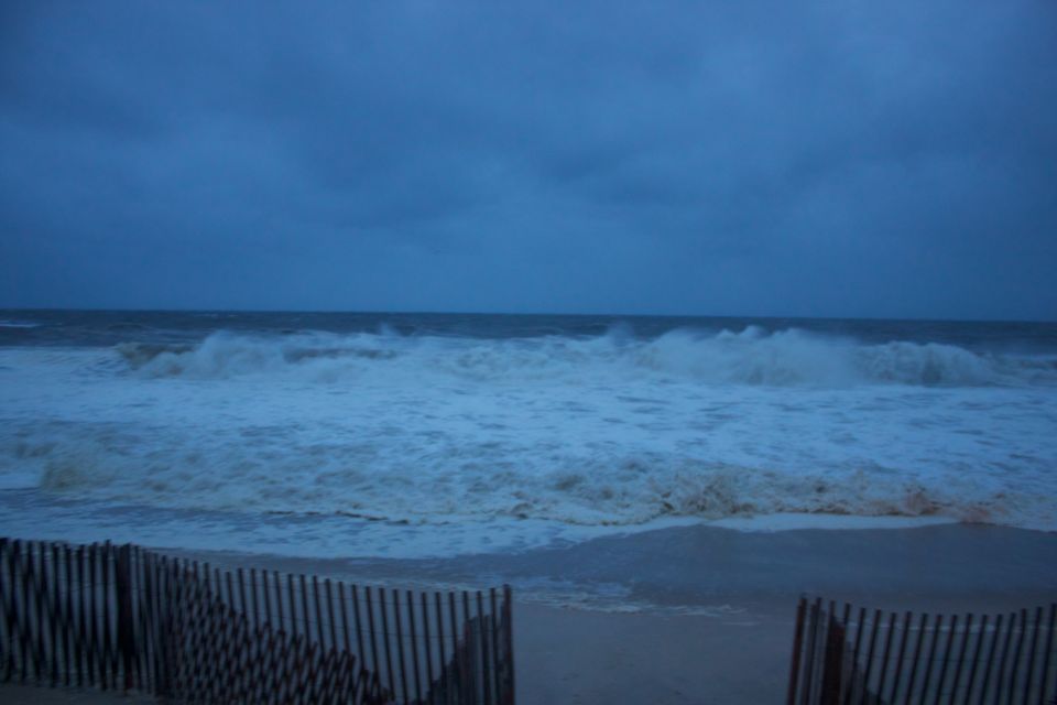 Stormy ocean waves crash on a beach with a wooden fence in the foreground. Supermotors.net logo visible